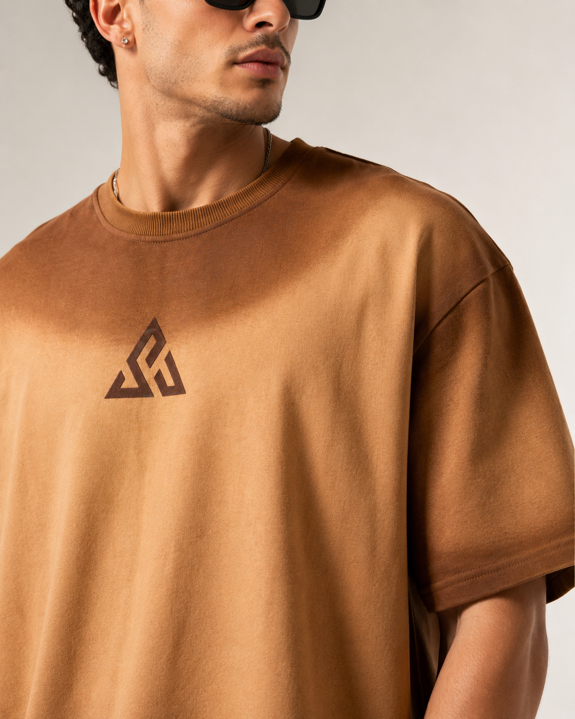A close-up of a man wearing a brown oversized crewneck t-shirt with a dark geometric logo on the chest, set against a plain light gray background.