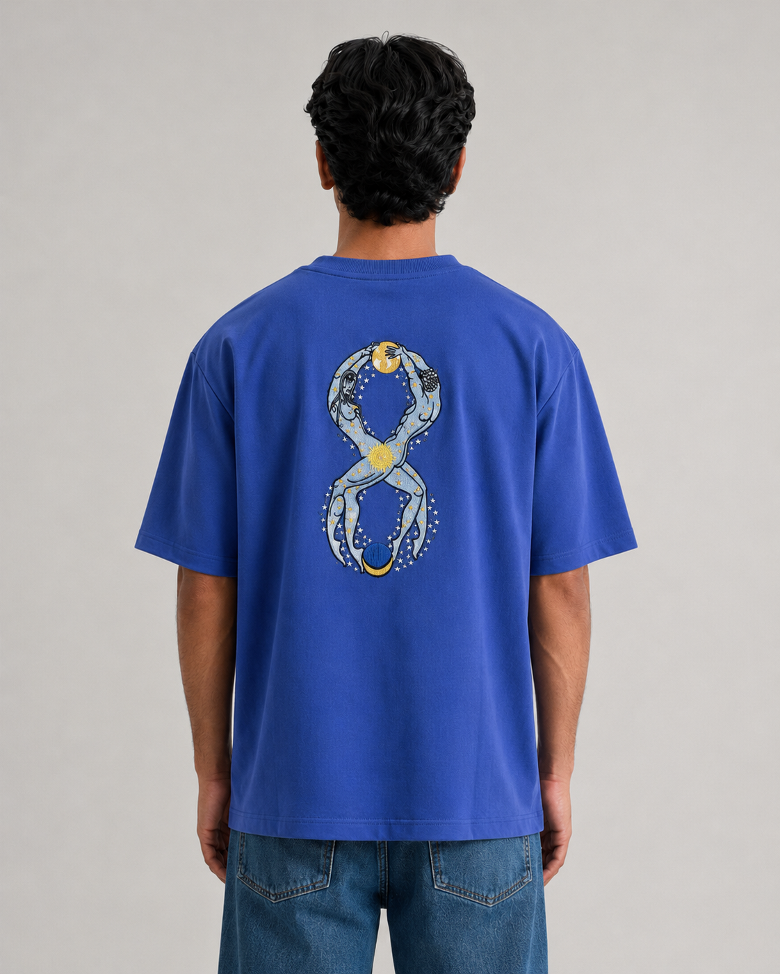 A man viewed from behind wearing a royal blue t-shirt with a large graphic print of a stylized infinity symbol on the back, paired with blue jeans against a neutral gray studio background.