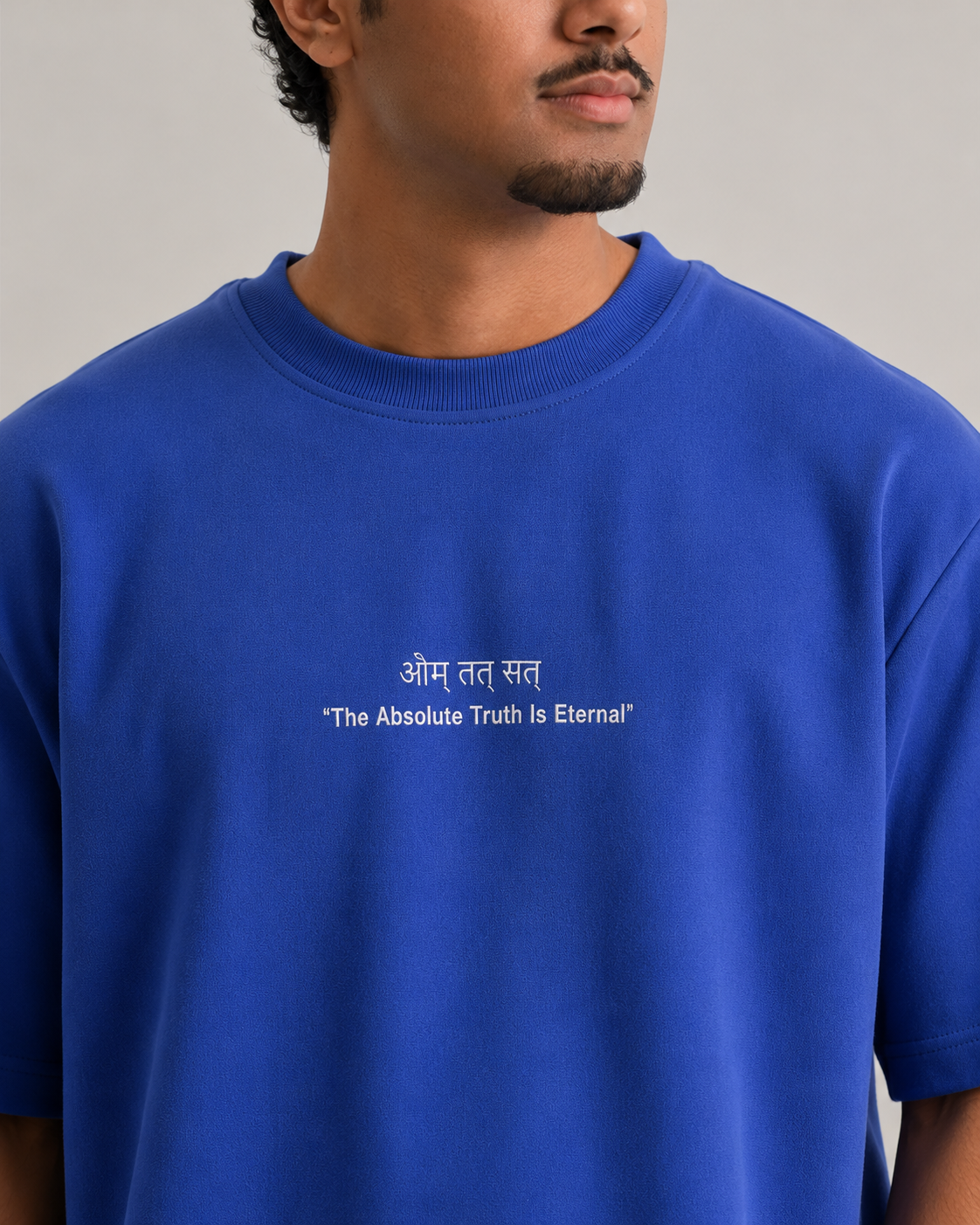 A close-up of a man wearing a royal blue crewneck sweatshirt with white printed text on the chest reading 'Om Tat Sat' and 'The Absolute Truth Is Eternal'.