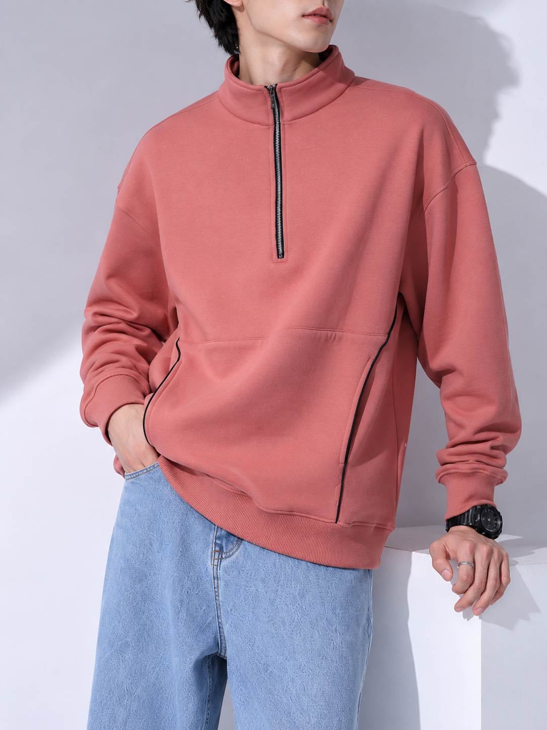 A man wearing a dusty rose quarter-zip pullover sweatshirt with black piping details and light blue jeans, standing against a white wall with soft shadows.