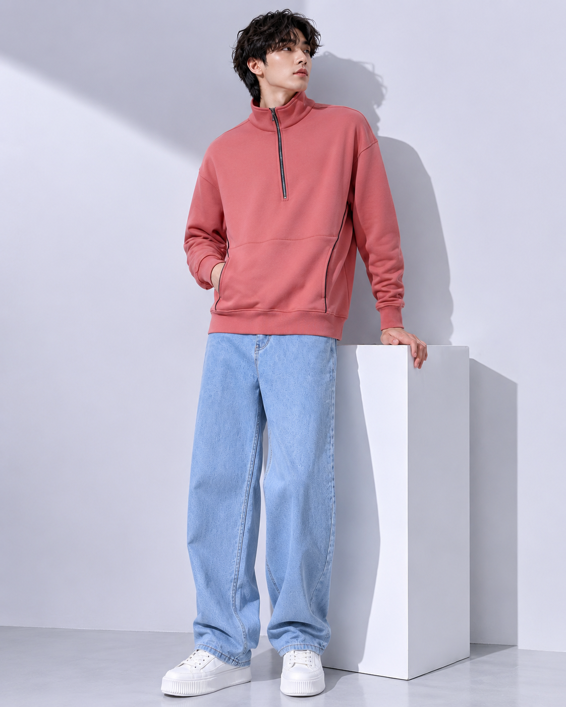 A model stands in a studio wearing a dusty rose quarter-zip pullover hoodie, light blue wide-leg jeans, and white sneakers, leaning against a white display block.