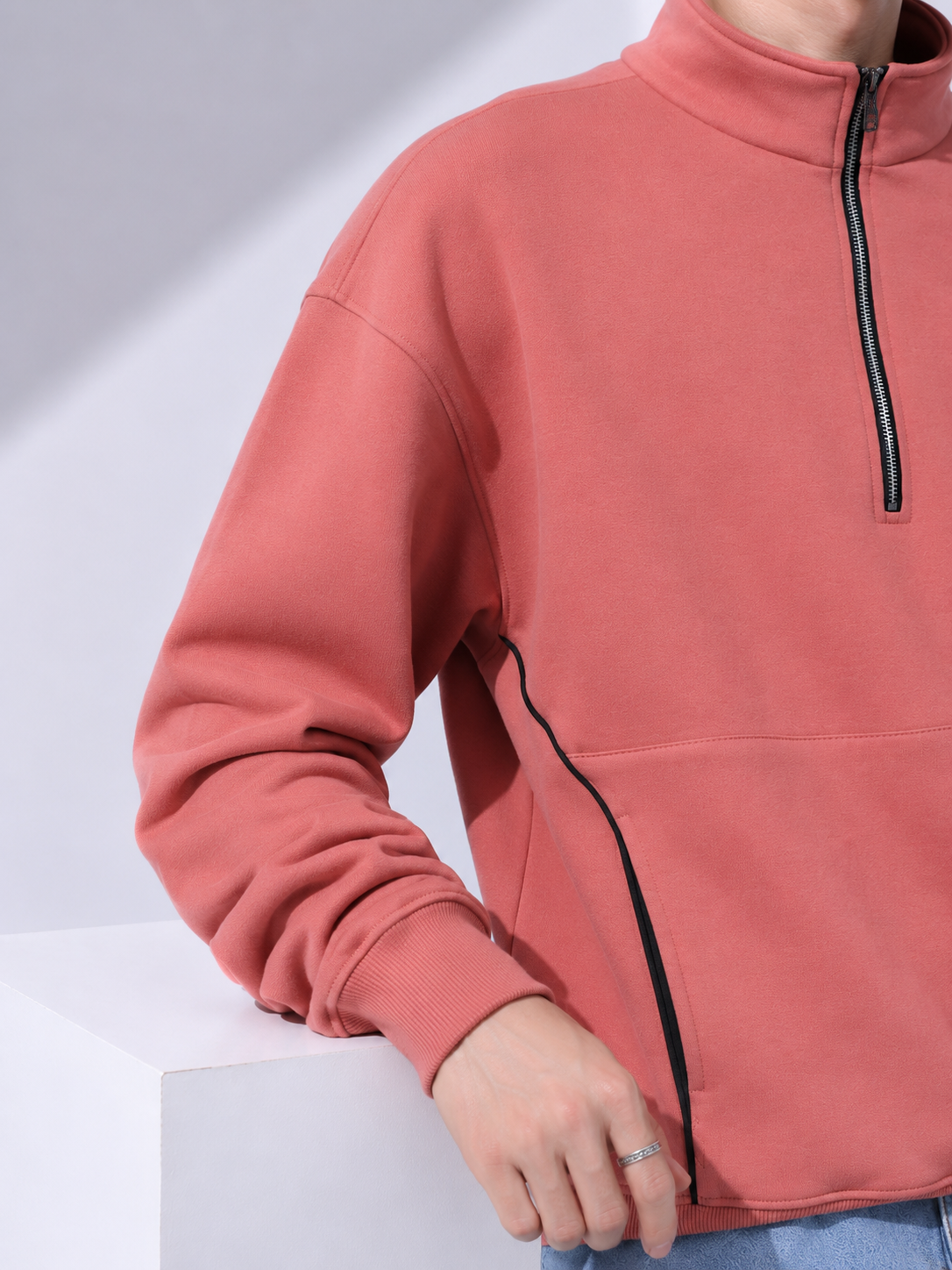 A close-up of a person wearing a coral pink quarter-zip sweatshirt with black piping details, resting on a white display block.