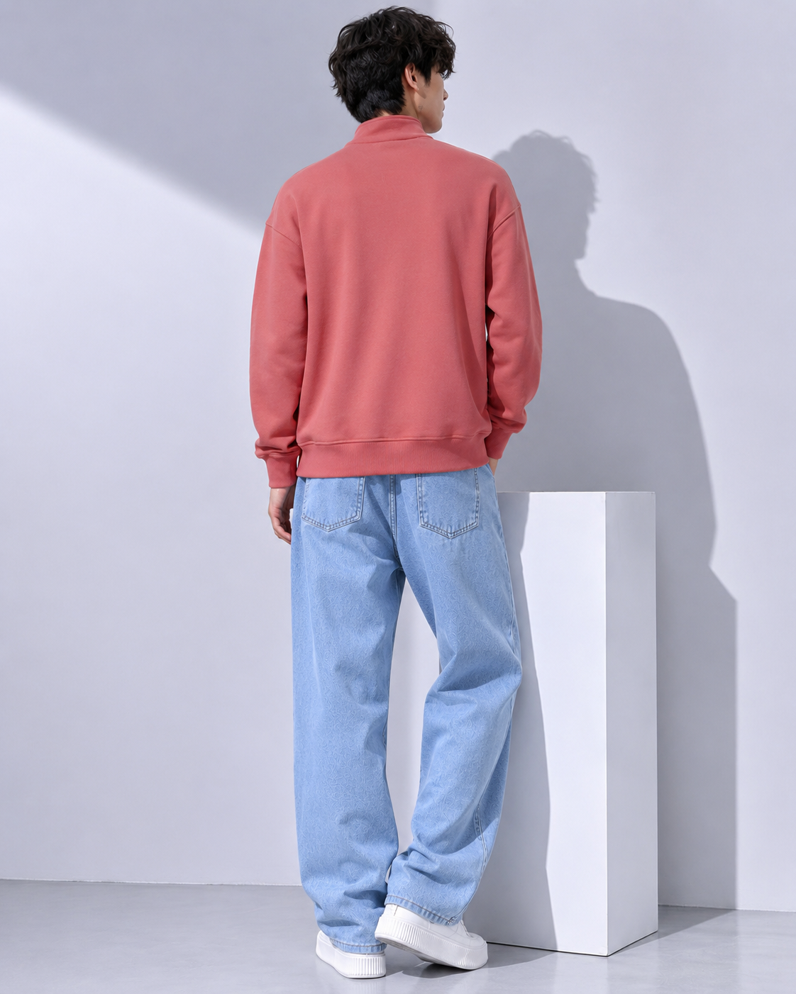 A man shown from the back wearing a coral pink sweatshirt and light blue jeans, standing next to a white display block in a minimalist studio setting.
