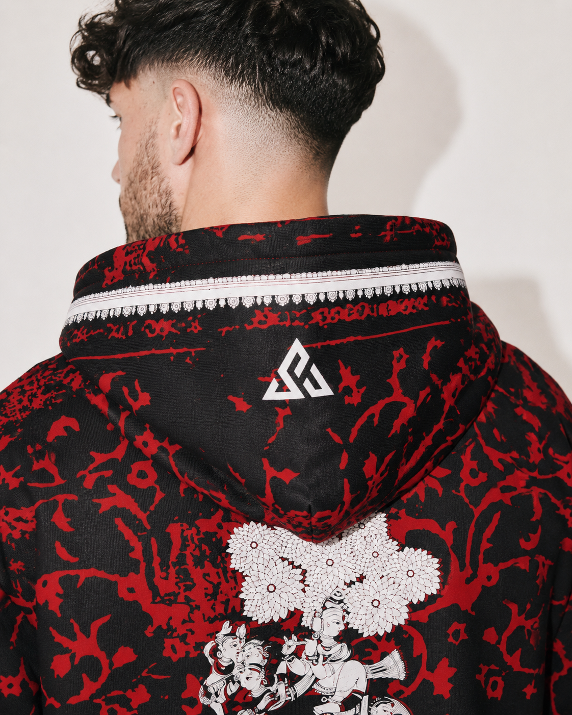 Close-up back view of a person wearing a black hooded jacket with a bold red abstract print, a white patterned collar trim, and a white geometric logo on the back of the neck.