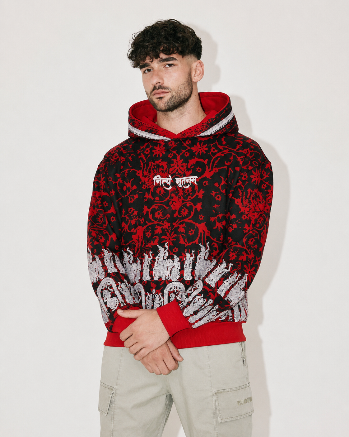 A man wearing a red and black patterned hoodie with white script on the chest and white sleeve graphics, paired with light khaki cargo pants, standing against a plain off-white background.
