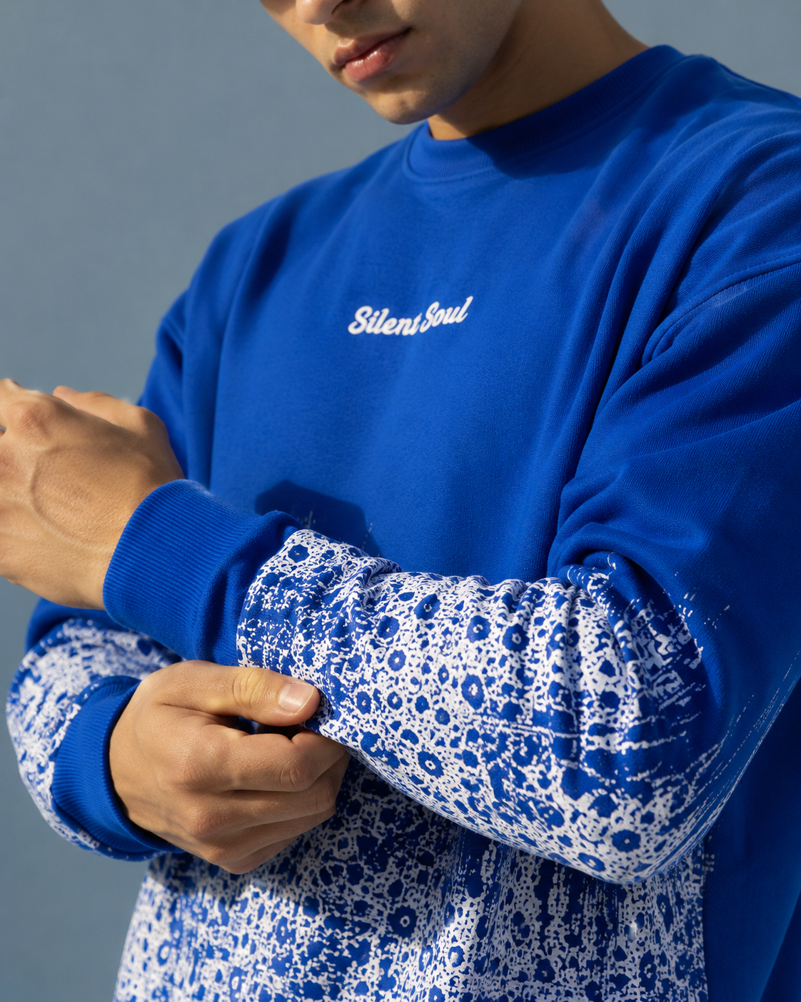 A close-up of a person wearing a royal blue crewneck sweatshirt with 'Silent Soul' embroidered in white on the chest. The sweatshirt features a blue and white splatter pattern on the lower body and cuffs, and is shown being adjusted on the arm.