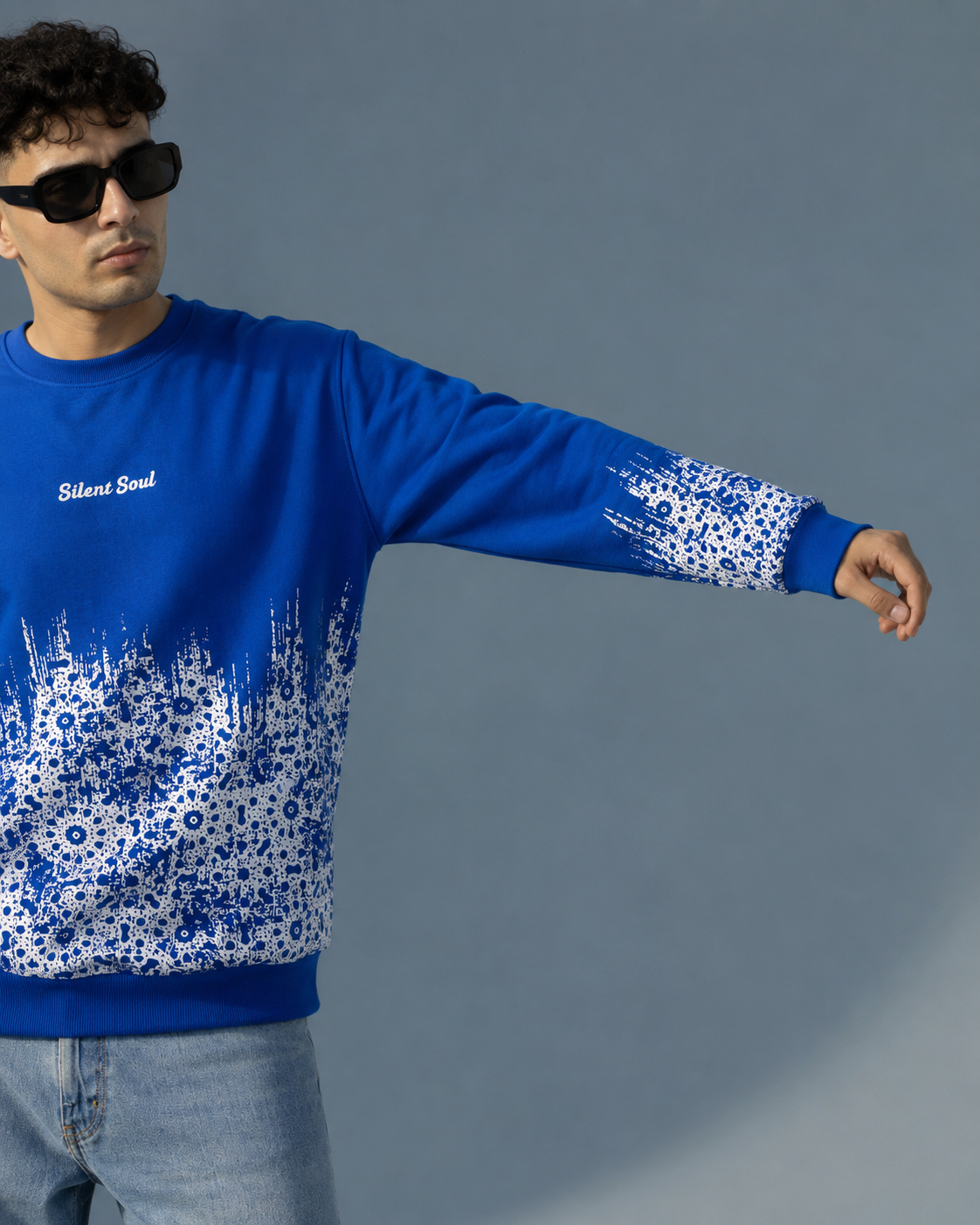 A man wearing a bright blue crewneck sweatshirt with a white floral pattern fading from the hem and cuffs, featuring the text 'Silk Soul' on the chest, paired with light blue jeans against a solid gray background.