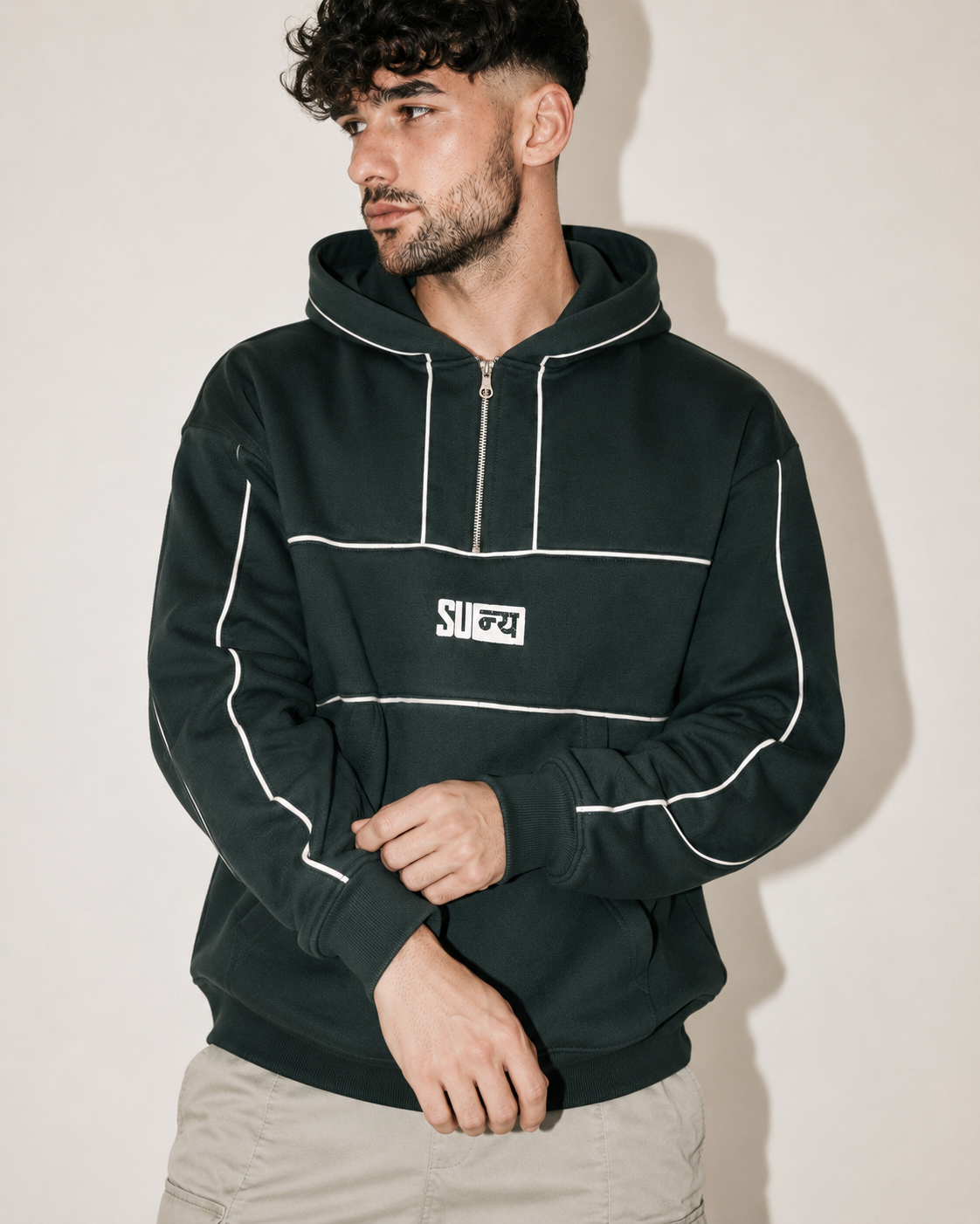 A man wearing a dark forest green half-zip hoodie with white contrast piping and a white chest logo, paired with light beige pants, set against a plain beige wall.