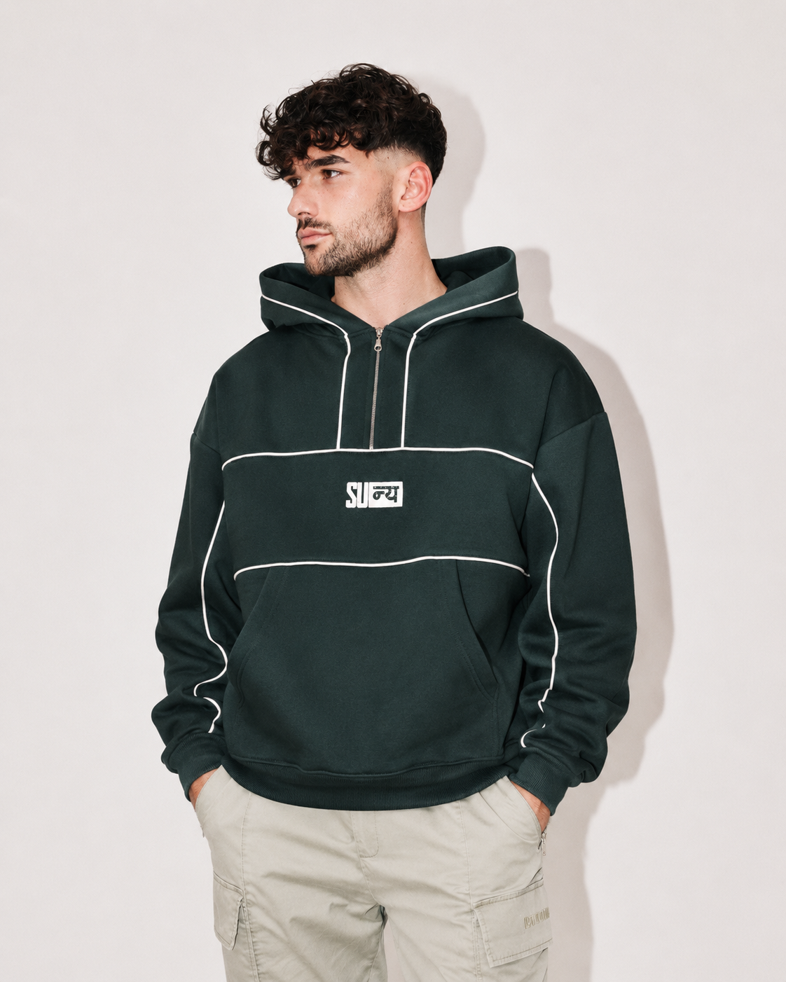 A man wearing a dark forest green pullover hoodie with white piping and a white chest logo, paired with light beige cargo pants, standing against a plain off-white background.