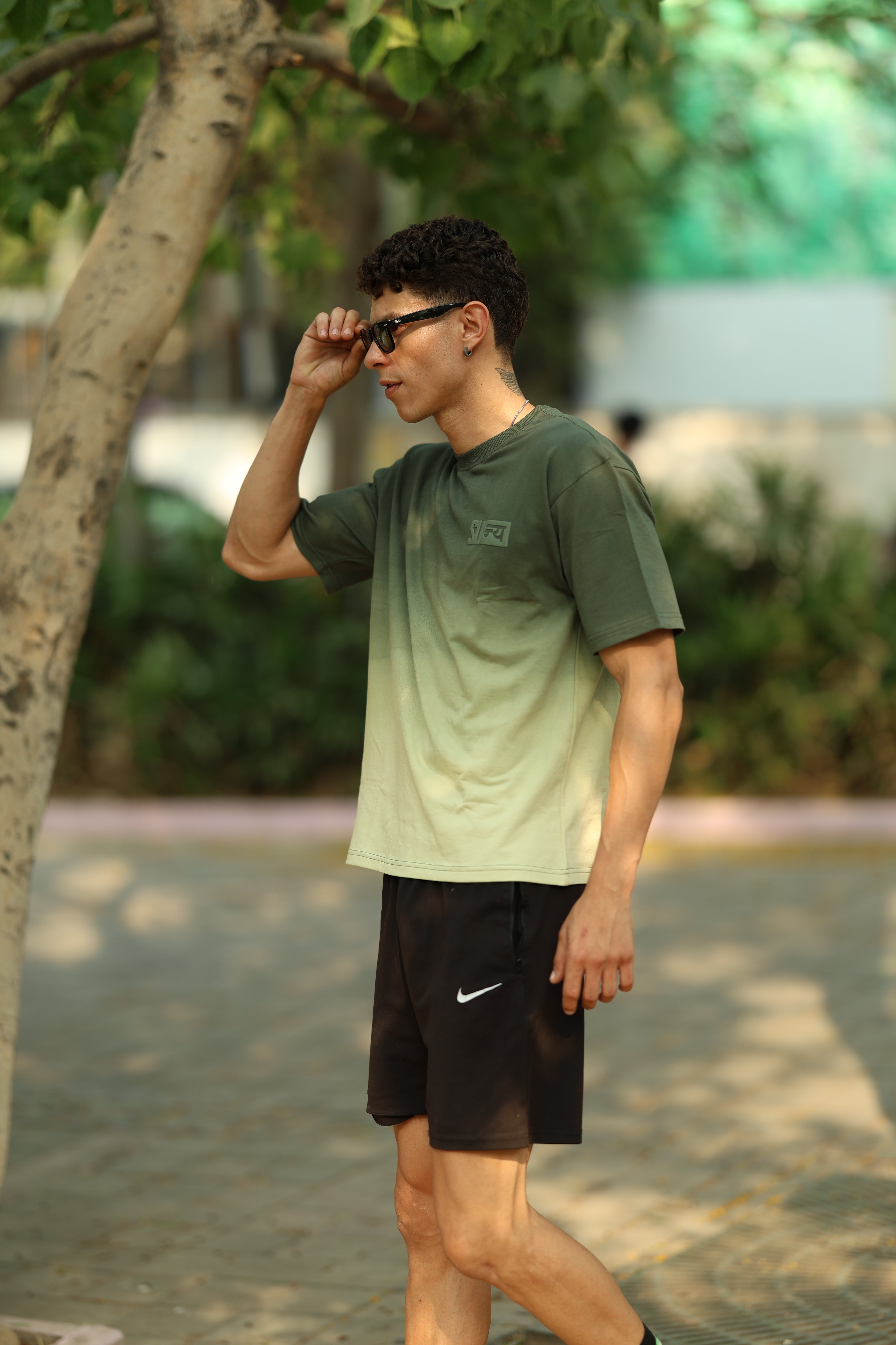 Ombrix tee in olive fading to sage — worn outdoors under trees, styled with black shorts and sunglasses