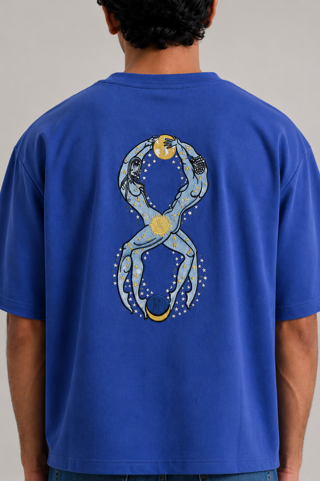 A man viewed from behind wearing a royal blue t-shirt with a large graphic print of two figures forming an infinity symbol, holding a crescent moon and star, on the back.