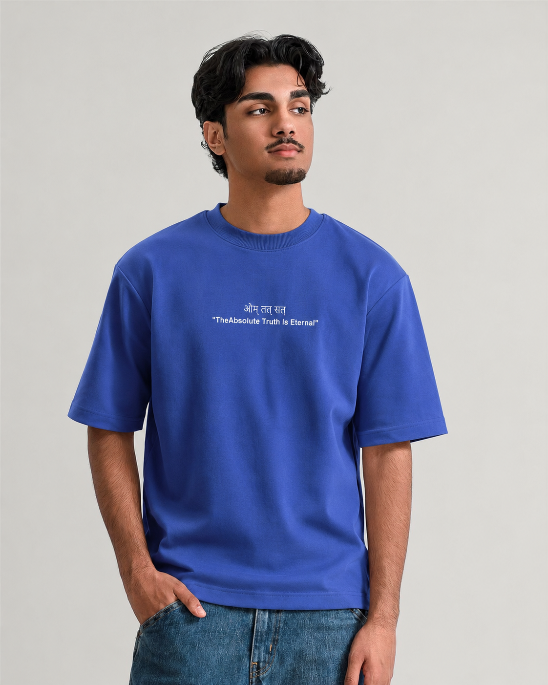 A man wearing a royal blue crewneck t-shirt with white printed text on the chest, paired with blue jeans, against a solid light gray background.