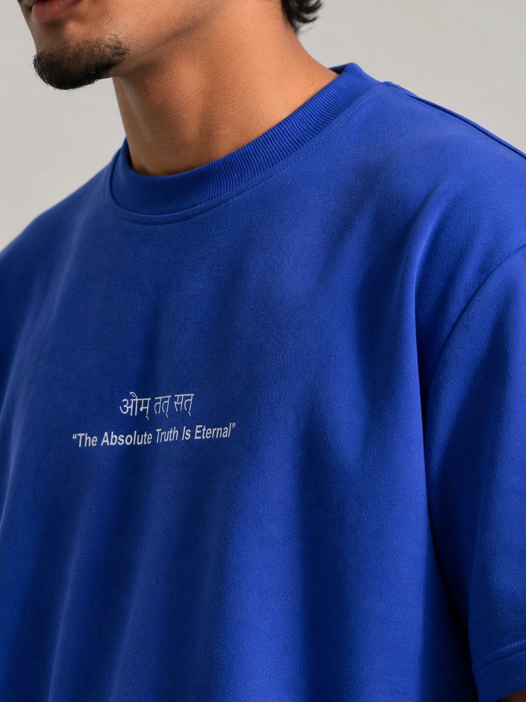 A close-up of a man wearing a royal blue crewneck t-shirt with white printed text on the chest reading 'Aum Tat Sat The Absolute Truth Is Eternal'.