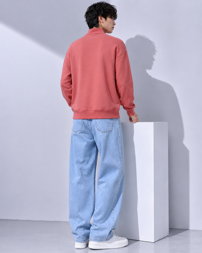 A man wearing a coral pink quarter-zip sweatshirt and light blue relaxed-fit jeans, shown from the back in a minimalist studio setting with a white pedestal.