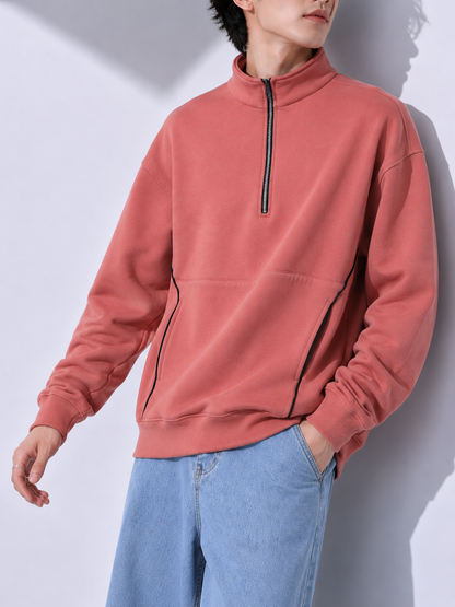 A man wearing a dusty rose quarter-zip sweatshirt with black piping details and light blue jeans, standing against a light gray wall with soft shadows.