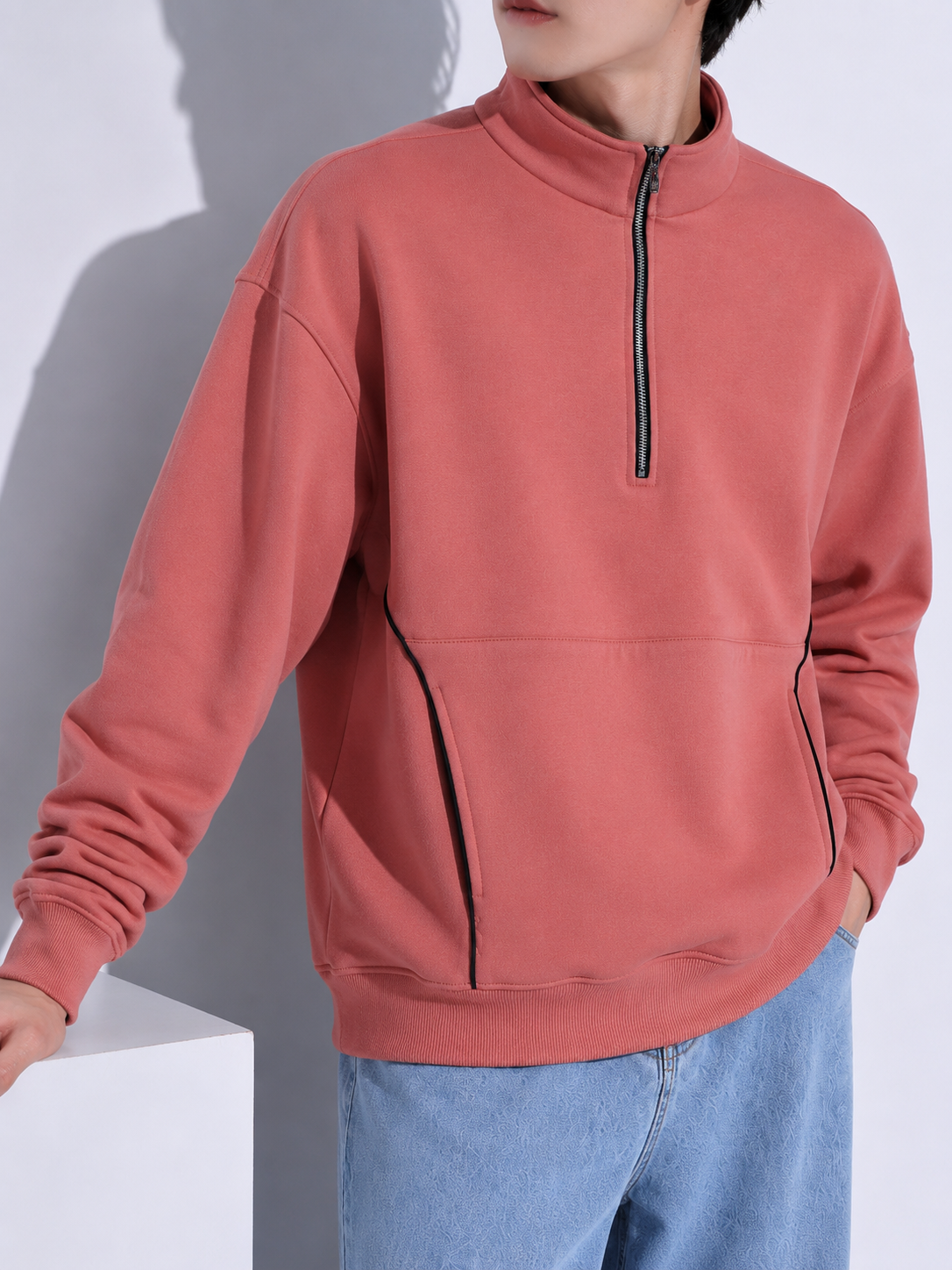 A person wearing a dusty rose quarter-zip sweatshirt with black piping details, paired with light blue jeans, shown against a gray background.