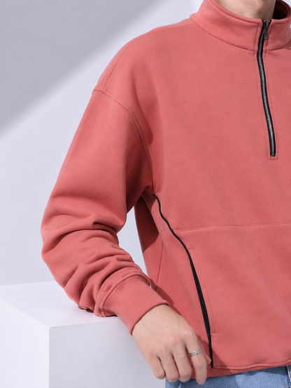 A close-up of a person wearing a coral pink quarter-zip sweatshirt with black piping details, resting on a white display block.