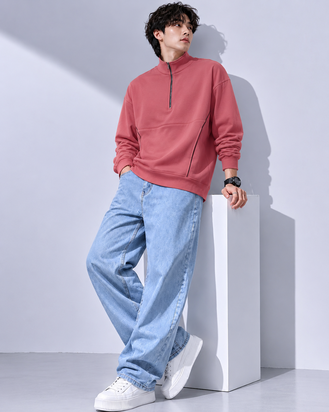 A man wearing a dusty rose quarter-zip sweatshirt, light blue wide-leg jeans, and white sneakers, leaning against a white block in a studio setting.