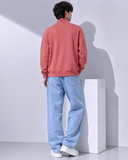 A man shown from the back wearing a coral pink sweatshirt and light blue jeans, standing next to a white display block in a minimalist studio setting.