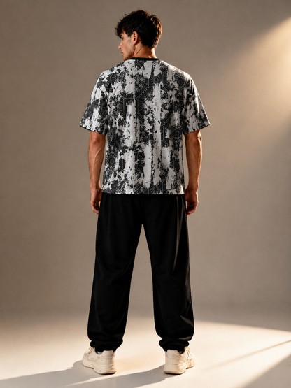 A man viewed from behind wearing a short-sleeve t-shirt with a black and white abstract pattern, black sweatpants, and white sneakers, standing against a warm-toned studio backdrop.
