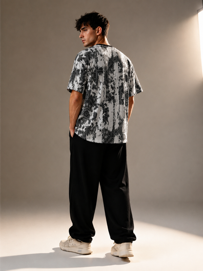 A man standing in a studio wearing a black and white abstract patterned t-shirt, loose black sweatpants, and white sneakers, shown from a rear three-quarter angle.
