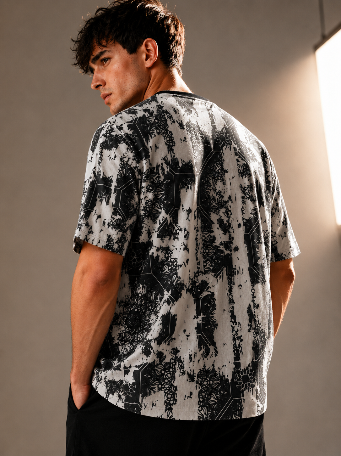 A man wearing a short-sleeve t-shirt with a black and white abstract geometric pattern, shown from a three-quarters back view in a studio setting.