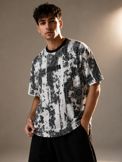 A man wearing a black and white abstract patterned t-shirt with a black crew neck, paired with black shorts, standing against a warm beige studio background.