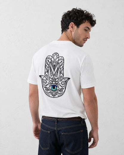 A man wearing a white t-shirt with a large black and blue Hamsa hand graphic on the back, paired with dark blue jeans, photographed against a light gray textured wall.