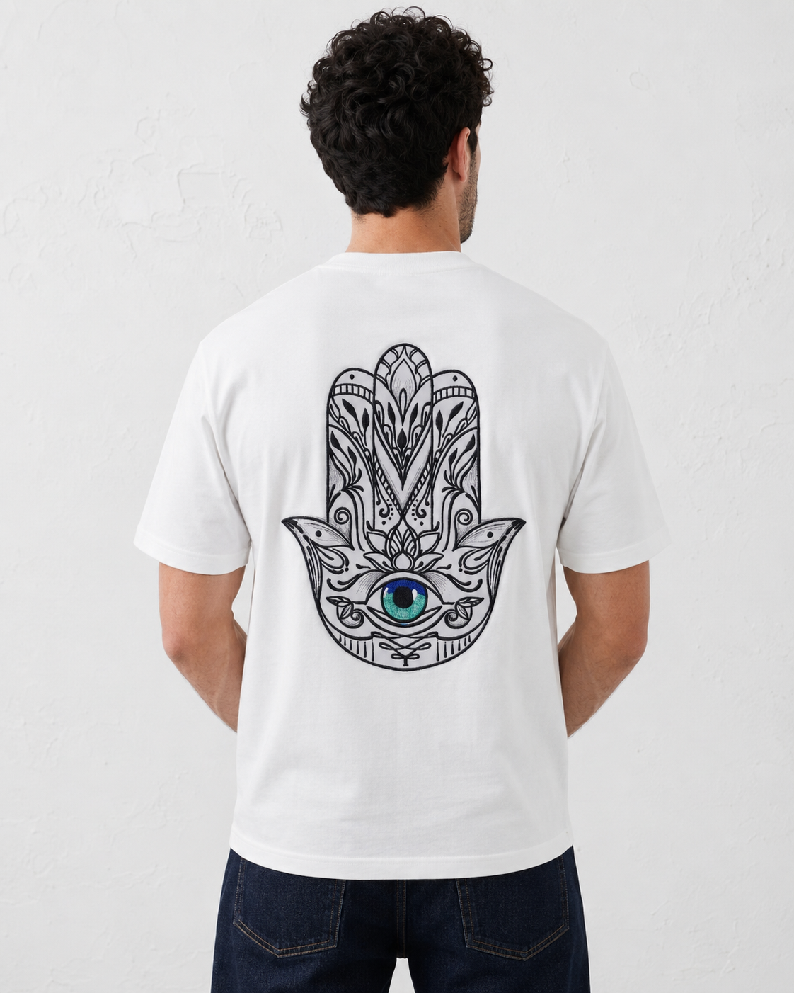 A man viewed from behind wearing a white t-shirt with a large black hamsa hand graphic on the back featuring a blue and green eye, paired with dark blue jeans against a light gray wall.