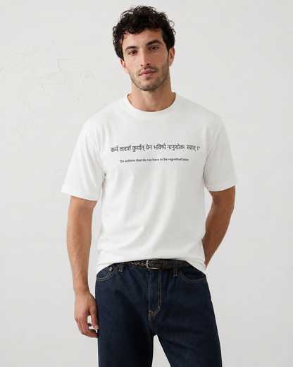 A man wearing a white short-sleeve crewneck t-shirt with black printed text in Devanagari script and English translation on the chest, paired with dark blue jeans, against a light gray textured wall.