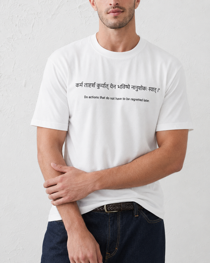 A man wearing a white short-sleeve crewneck t-shirt with black printed text in Devanagari script and English on the chest, paired with dark blue jeans against a textured white wall.