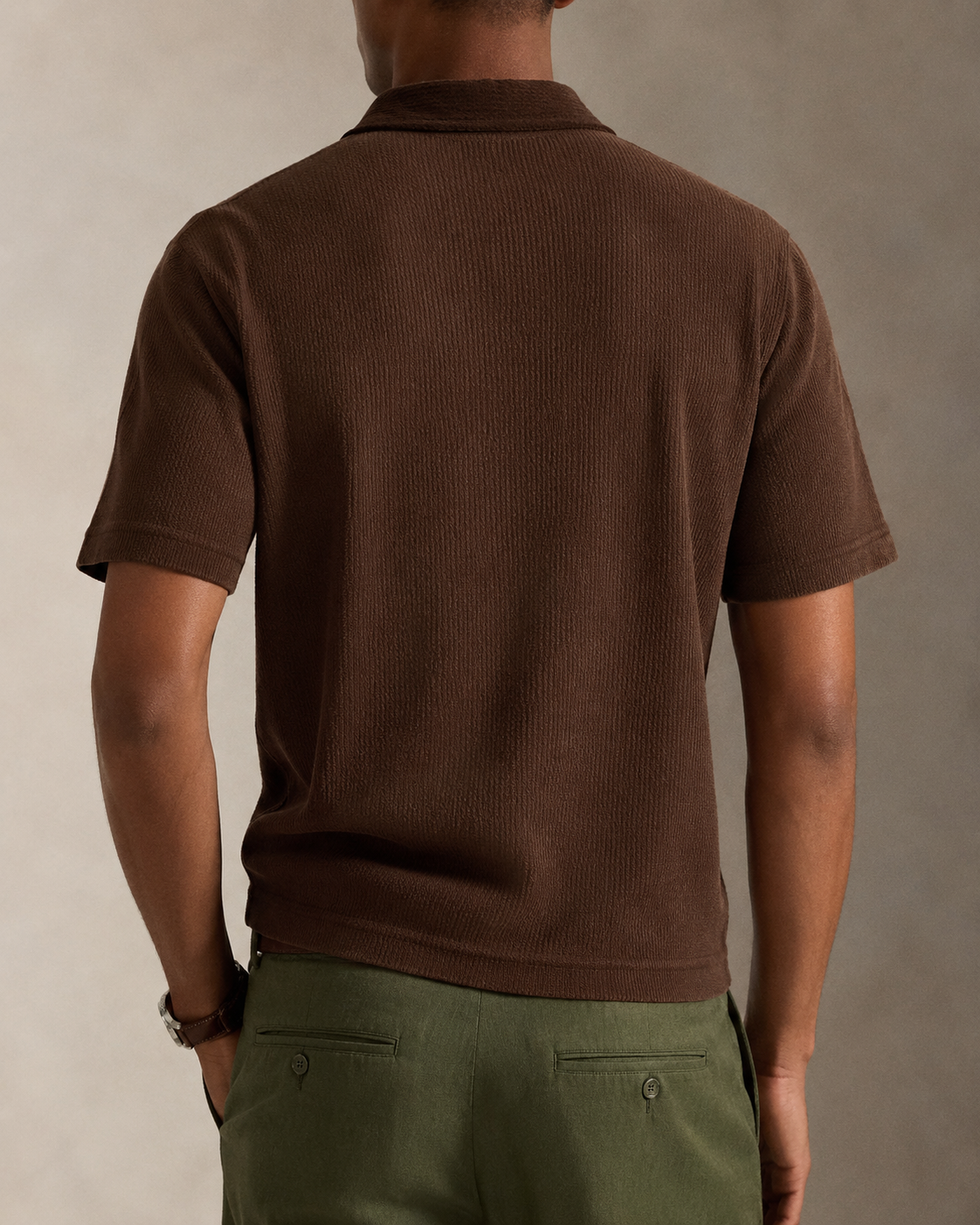 A man viewed from behind wearing a brown short-sleeve ribbed knit polo shirt and olive green trousers, set against a neutral studio background.