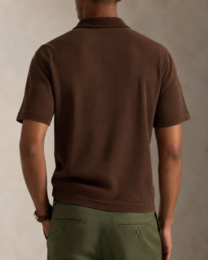 A man viewed from behind wearing a brown short-sleeve ribbed knit polo shirt and olive green trousers, set against a neutral studio background.