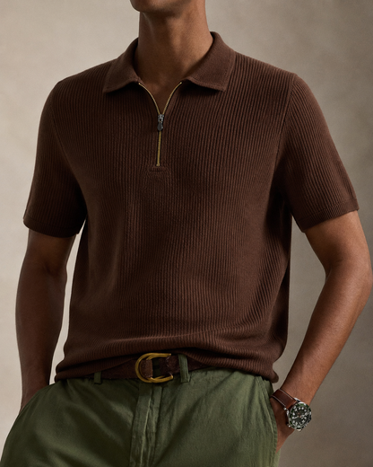 A man wearing a brown ribbed short-sleeve polo shirt with a half-zip collar, styled with olive green trousers and a gold-buckled belt against a warm beige studio background.