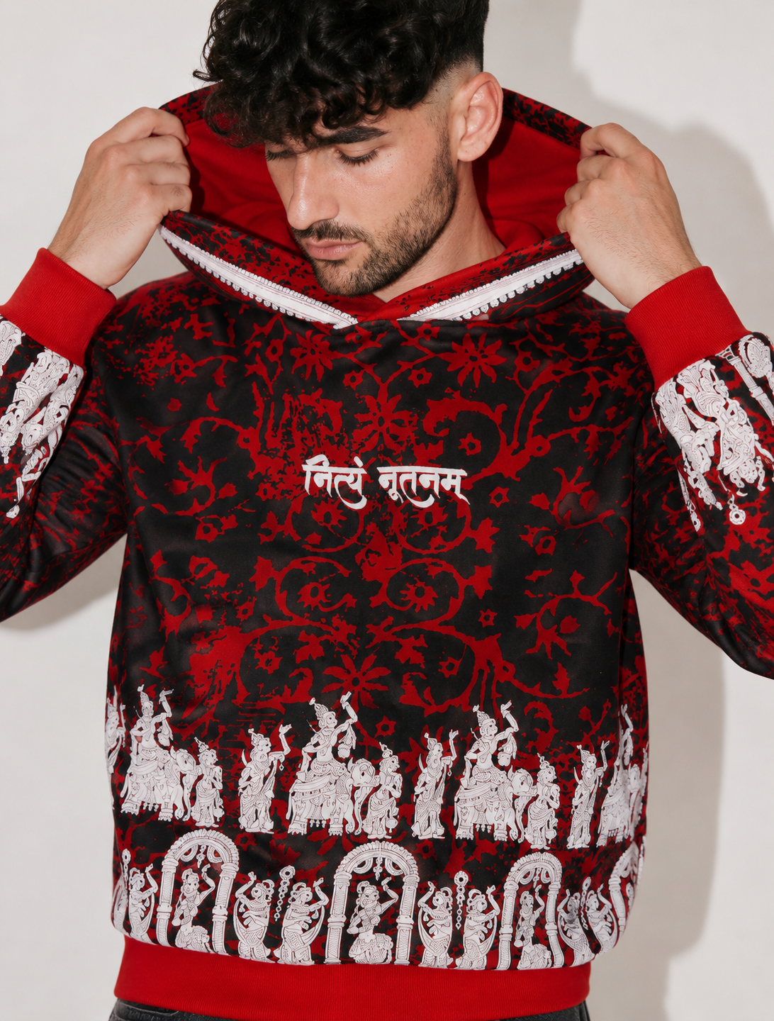 A man wearing a black and red patterned hoodie with white elephant motifs and Hindi text on the chest, holding up the hood.