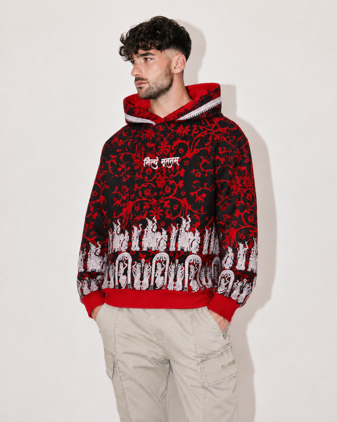 A man wearing a red and black patterned hoodie with white graphic details on the sleeves and chest, paired with light beige cargo pants, standing against a plain light gray background.