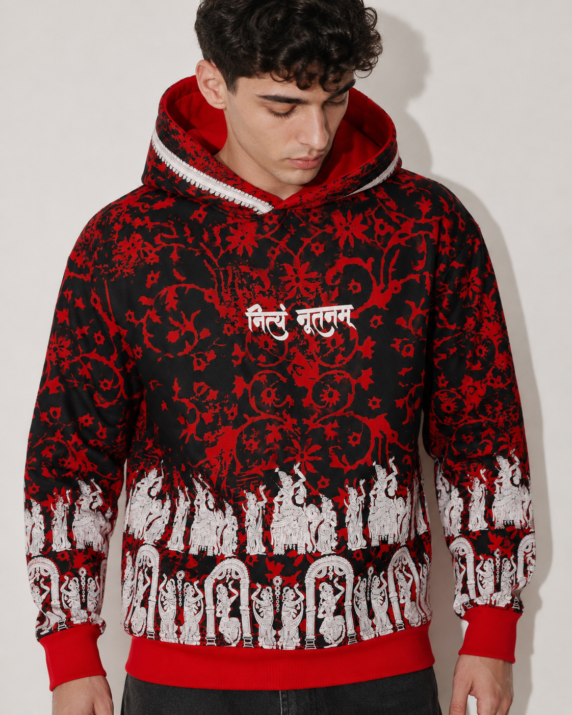 A man wearing a black and red patterned hoodie with white script on the chest and a white graphic border along the hem, photographed against a plain light background.