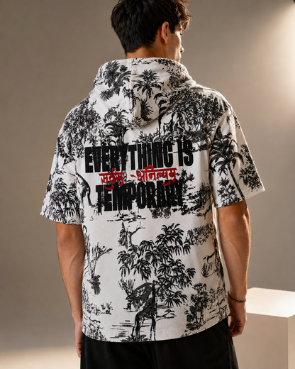 A man viewed from behind wearing a white short-sleeve hooded shirt with a black tropical tree print and bold text across the back reading 'EVERYTHING IS TEMPORARY' with red script overlay, against a warm beige studio background.