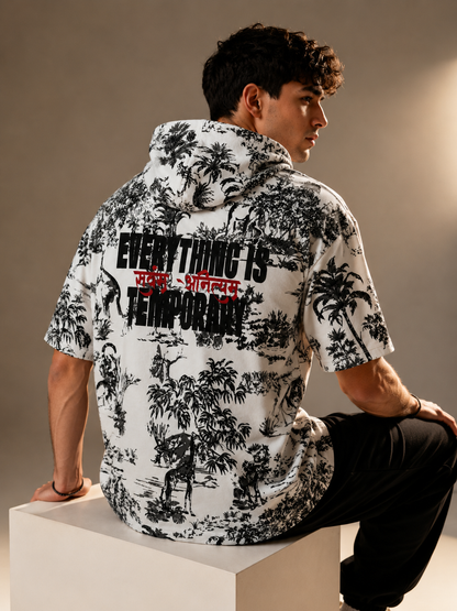 A man wearing a white short-sleeve button-up shirt with a black tropical print and bold text on the back, seated on a white block against a warm beige studio background.