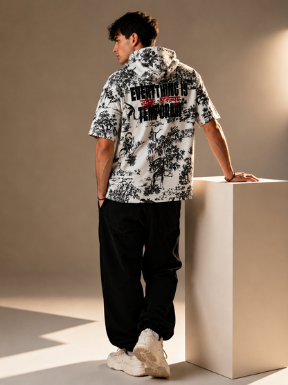 A man stands on a beige pedestal wearing a white short-sleeve hooded shirt with a black tropical tree print and bold black text on the back, paired with black pants and white sneakers in a studio setting.