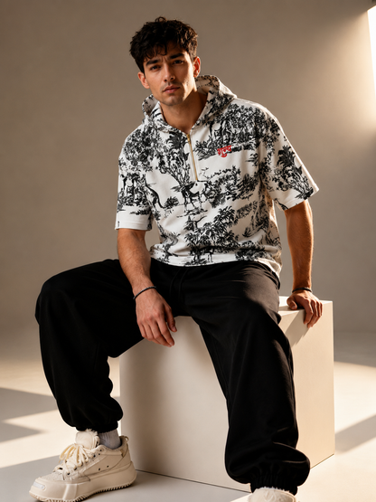 A man wearing a white short-sleeve quarter-zip hoodie with a black toile de Jouy pattern and a small red embroidered logo on the chest, paired with black sweatpants and off-white sneakers, sitting on a white block in a studio setting.
