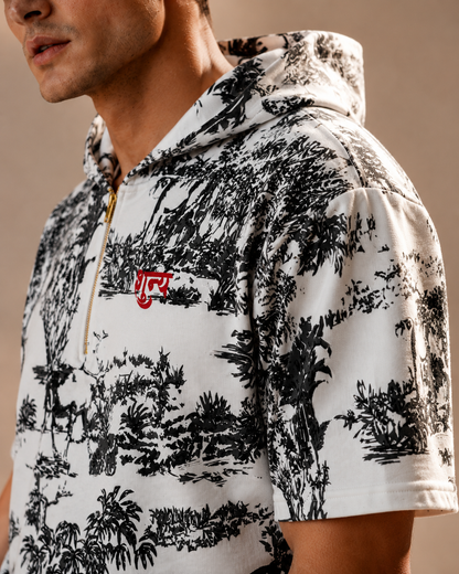 A close-up of a man wearing a white short-sleeve half-zip hoodie with a black toile de Jouy landscape print and a red embroidered chest logo.