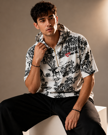 A man wearing a white short-sleeve hooded shirt with a black toile-style print and a small red embroidered logo on the chest, paired with black pants, seated on a white block against a warm beige studio background.