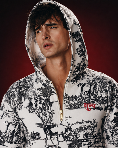 A man wearing a white zip-up hoodie with a black toile-style forest and deer print, featuring a red embroidered logo on the chest, set against a dark red studio background.