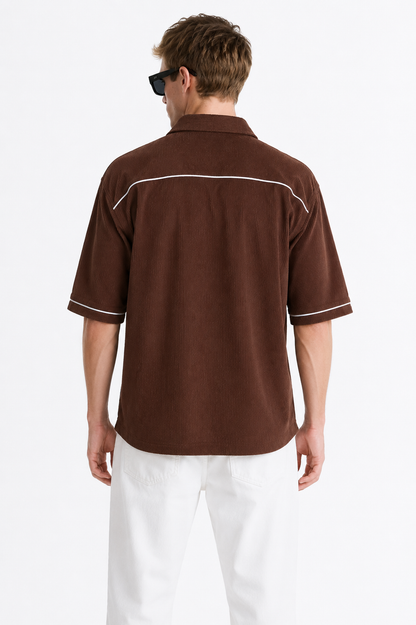 A man viewed from the back wearing a brown short-sleeve button-up shirt with white contrast piping and white pants, against a plain light background.