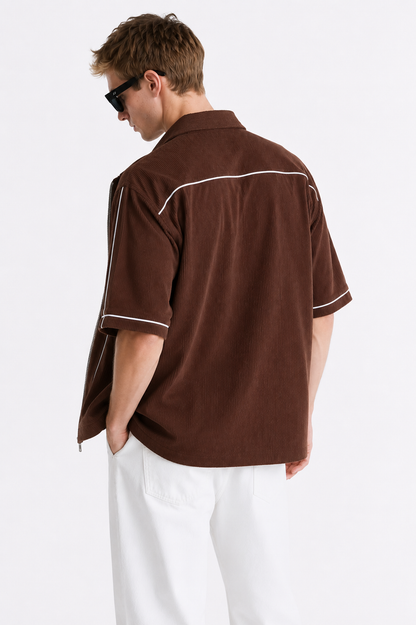 A man wearing a brown short-sleeve button-up shirt with white contrast piping, shown from the back against a plain white studio background.