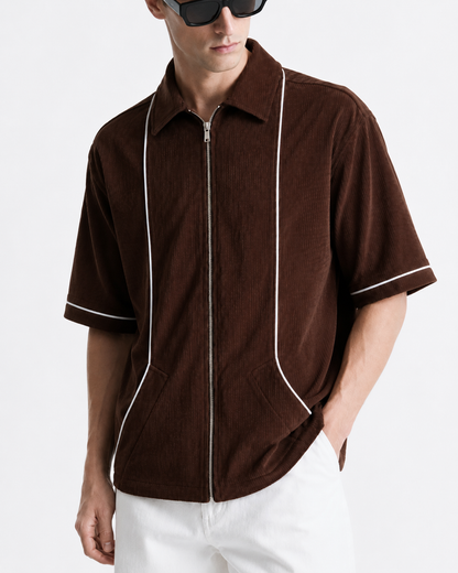 A man wearing a brown corduroy short-sleeve zip-up shirt with white piping details, paired with white pants and black sunglasses, against a plain white background.