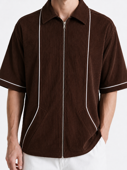 A man wearing a brown corduroy short-sleeve zip-up shirt with white piping details, paired with white pants, against a plain white background.