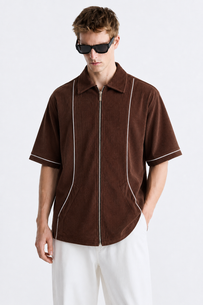 A man wearing a brown corduroy short-sleeve shirt with a front zipper and white piping details, paired with white pants and black sunglasses, against a plain light gray background.