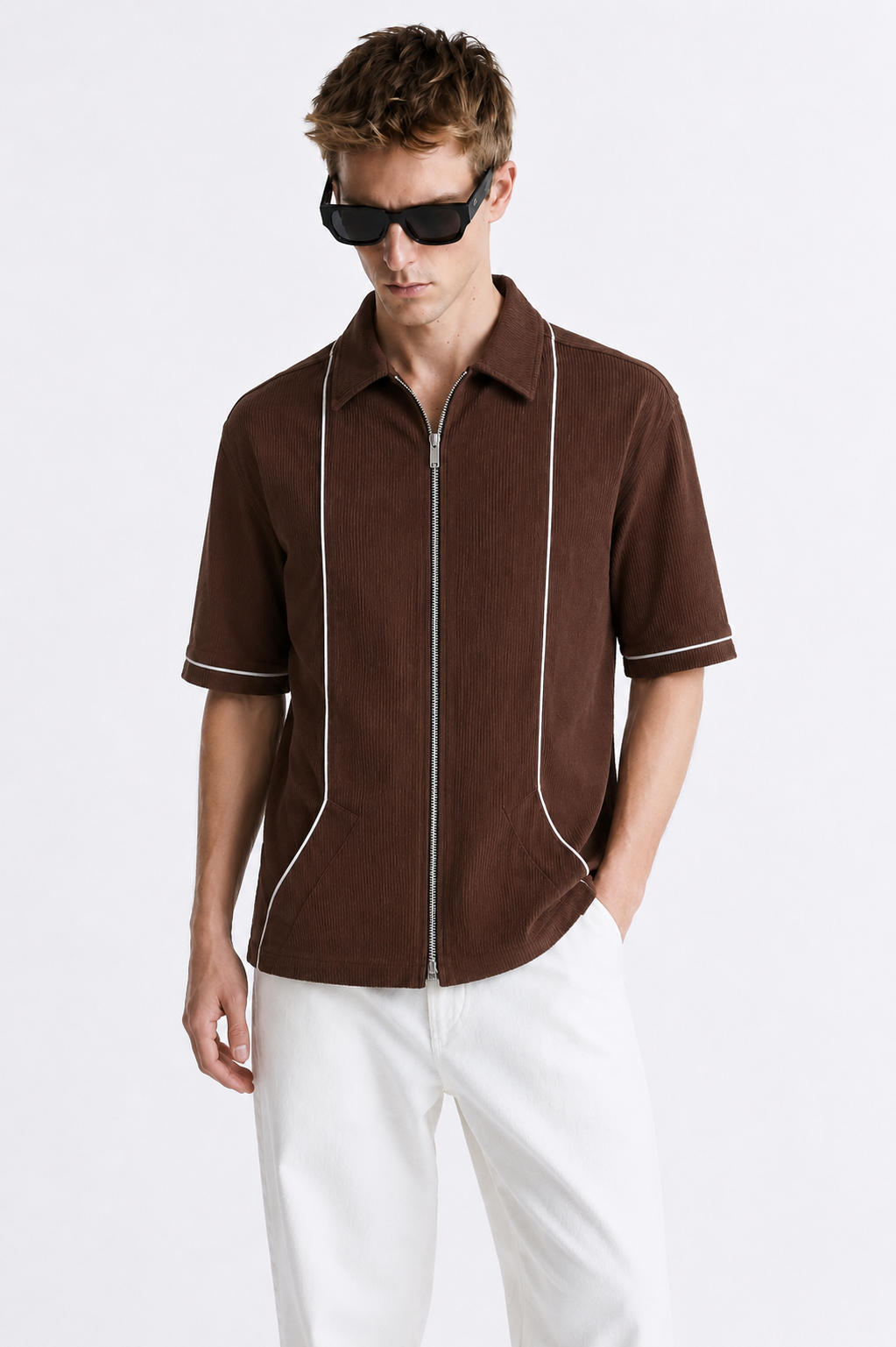A man wearing a brown short-sleeve zip-front shirt with white piping details, paired with white trousers and black sunglasses, against a plain white studio background.